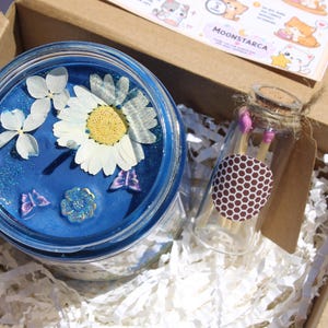 May include: A blue candle in a jar with dried flowers and butterfly accents, presented in a cardboard box with a small glass bottle of matches. The candle has a floral design.