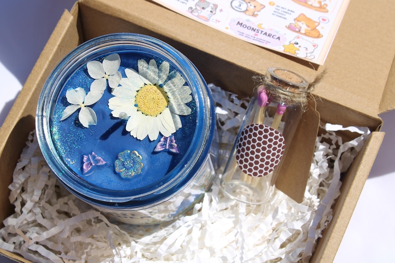 May include: A blue candle in a jar with dried flowers and butterfly accents, presented in a cardboard box with a small glass bottle of matches. The candle has a floral design.