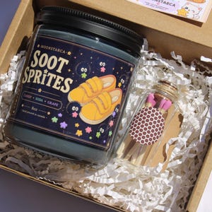 May include: A gift box containing a dark blue candle jar with the text "Soot Sprites" and a graphic of yellow shoes. A small glass jar of matches with pink tips and a brown polka dot label is also in the box.