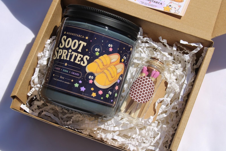 May include: A gift box containing a dark blue candle jar with the text "Soot Sprites" and a graphic of yellow shoes. A small glass jar of matches with pink tips and a brown polka dot label is also in the box.