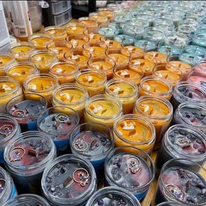 May include: A collection of scented candles in glass jars, arranged in rows. The candles feature various colors, including orange, blue, and purple. Some candles have decorative elements like crystals and charms. The candles are on a wooden surface.