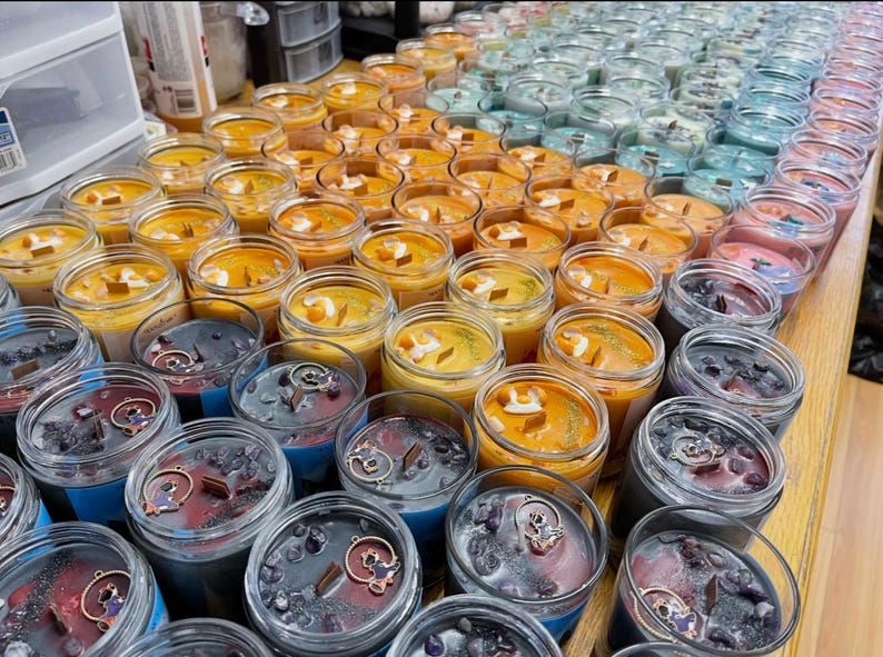 May include: A collection of scented candles in glass jars, arranged in rows. The candles feature various colors, including orange, blue, and purple. Some candles have decorative elements like crystals and charms. The candles are on a wooden surface.