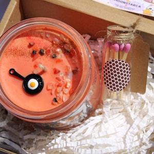 May include: A candle in a glass jar with an orange wax, decorated with small stones and a black and white fried egg charm. A small glass bottle of matches is included in the box, along with white paper filler.