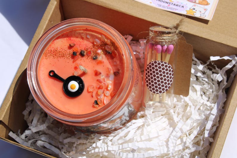 May include: A candle in a glass jar with an orange wax, decorated with small stones and a black and white fried egg charm. A small glass bottle of matches is included in the box, along with white paper filler.