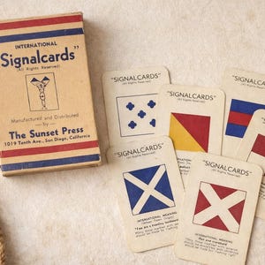 May include: Vintage "Signalcards" card game set. The box is tan with red and blue stripes and text. Several signal cards are displayed, each with a unique flag design in red, blue, and yellow. The cards are from The Sunset Press.