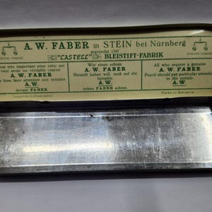 May include: A rectangular metal pencil case with a hinged lid. The interior of the lid features green text and graphics on a cream background, including the brand name A.W. Faber, the location Stein bei N&uuml;rnberg, and instructions in three languages.