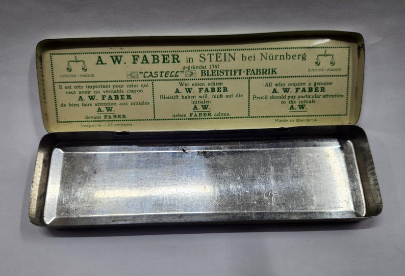 May include: A rectangular metal pencil case with a hinged lid. The interior of the lid features green text and graphics on a cream background, including the brand name A.W. Faber, the location Stein bei N&uuml;rnberg, and instructions in three languages.