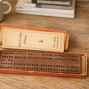 May include: A vintage cribbage board game with a wooden playing surface and numerous holes. The game is in an open orange box, with a booklet titled "How To Play Cribbage" visible. A white mug and a stack of books are in the background.