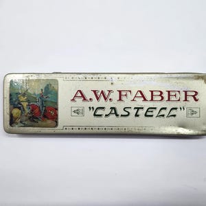 May include: A rectangular metal pencil case with a silver finish. The lid features a small illustration of a knight on horseback and the text A.W. FABER "CASTELL" in red and black lettering. The case shows signs of wear and age on the edges.