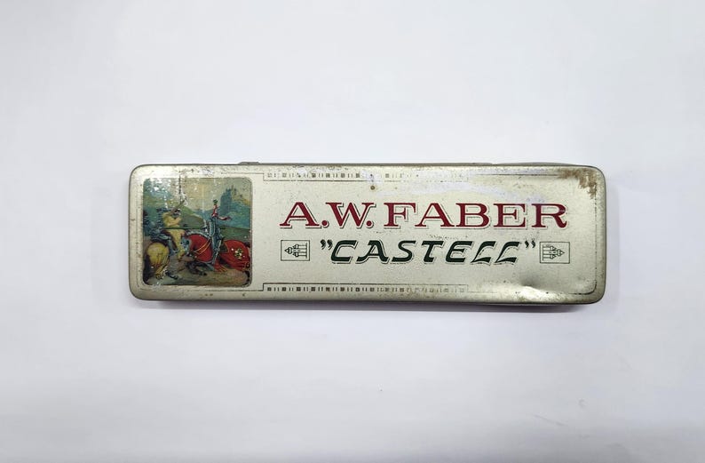 May include: A rectangular metal pencil case with a silver finish. The lid features a small illustration of a knight on horseback and the text A.W. FABER "CASTELL" in red and black lettering. The case shows signs of wear and age on the edges.