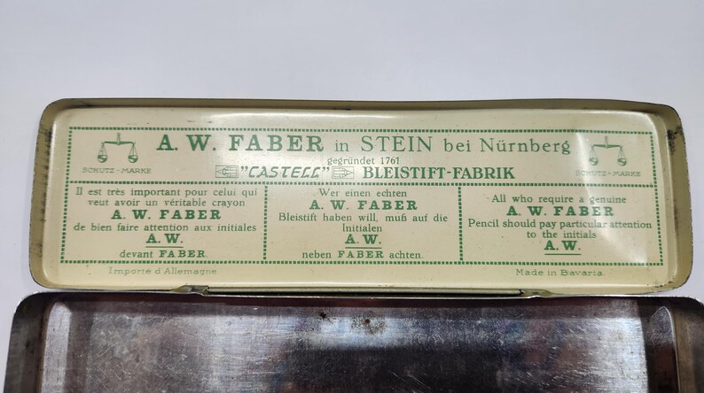 May include: A rectangular metal pencil tin with a cream-colored lid featuring green text and graphics. The lid displays the A.W. Faber brand name, the text "CASTELL" BLEISTIFT-FABRIK, and instructions in French, German, and English regarding the initials A.W.