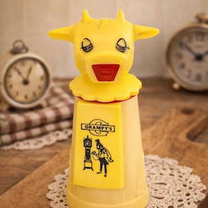 May include: A vintage yellow cow-shaped container with a red mouth and black eyes. The container has a yellow label with the text "Grampy's" and a black illustration of a man and a clock. The container is on a wooden cutting board.