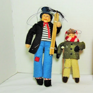 Puede incluir: Dos muñecos de tela hechos a mano, uno que representa a un deshollinador con abrigo negro, camisa a rayas y pantalones azules, y el otro un niño con abrigo de invierno y bufanda. Una lata roja de Coke Zero está al lado de los muñecos.