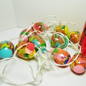 Peut inclure: Une guirlande de lumières en forme d'œufs de Pâques avec des illustrations colorées de lapins et d'autres thèmes de Pâques. Les lumières sont branchées sur une prise électrique et sont prêtes à être accrochées. Les lumières sont à côté d'une canette de Coca-Cola Zero Sugar.