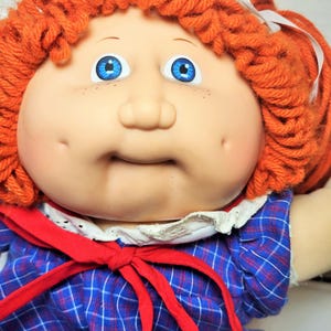 以下が含まれることがあります： 赤い毛糸の髪、青い目、青と白の格子縞のドレスに赤いリボンをつけたキャベツパッチキッズ人形です。
