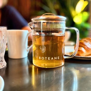 May include: A clear glass teapot with a stainless steel lid and a built-in strainer. The teapot is filled with a light brown tea and the word "BOTEANE" is printed on the side.