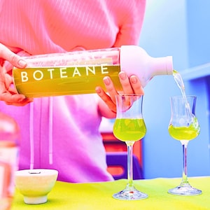Op de afbeelding: Een persoon giet een groene vloeistof uit een witte en doorzichtige fles met het label "BOTEANE" in twee heldere glazen shotglazen.