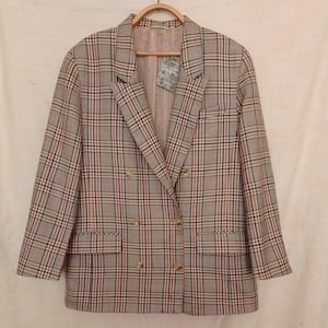Puede incluir: Un blazer cruzado con un estampado de cuadros en tonos beige, rojo y negro. El blazer tiene botones dorados y dos bolsillos delanteros. La chaqueta está colgada en una percha de madera.