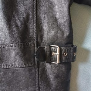 Pu&ograve; includere: Primo piano di una giacca di pelle nera. La giacca presenta una cintura con fibbia argentata e occhielli. La pelle ha un aspetto strutturato e le cuciture sono visibili lungo le cuciture. La giacca &egrave; uno stile classico.