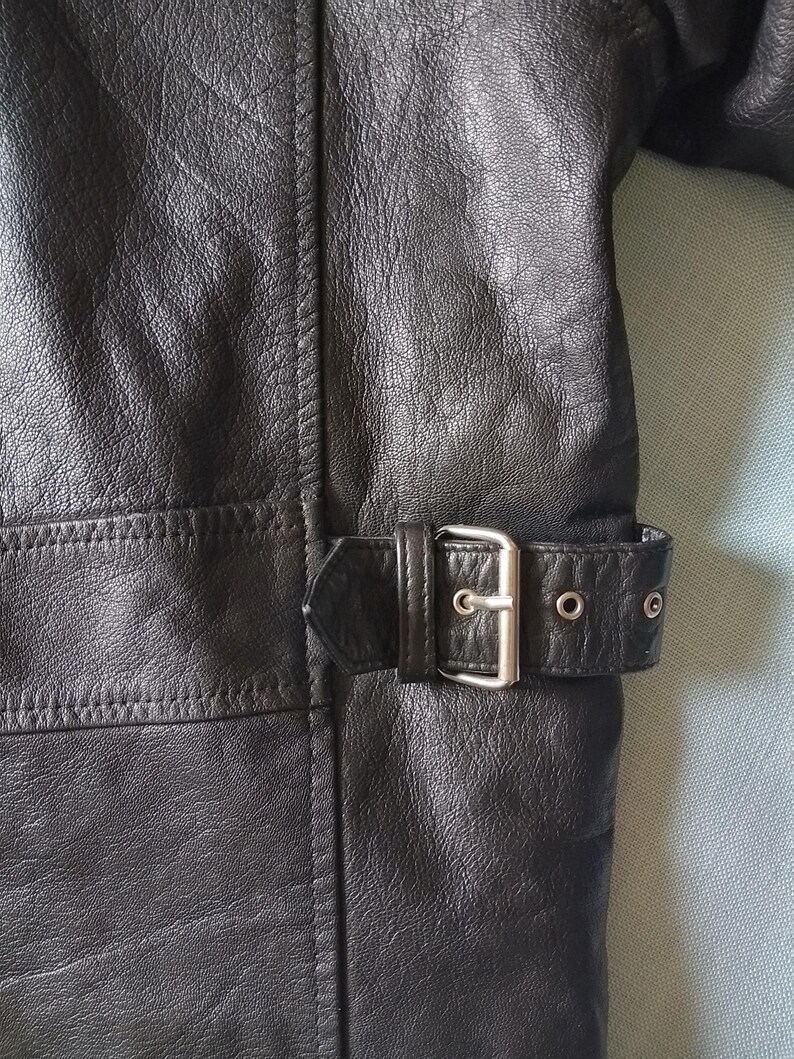 Pu&ograve; includere: Primo piano di una giacca di pelle nera. La giacca presenta una cintura con fibbia argentata e occhielli. La pelle ha un aspetto strutturato e le cuciture sono visibili lungo le cuciture. La giacca &egrave; uno stile classico.