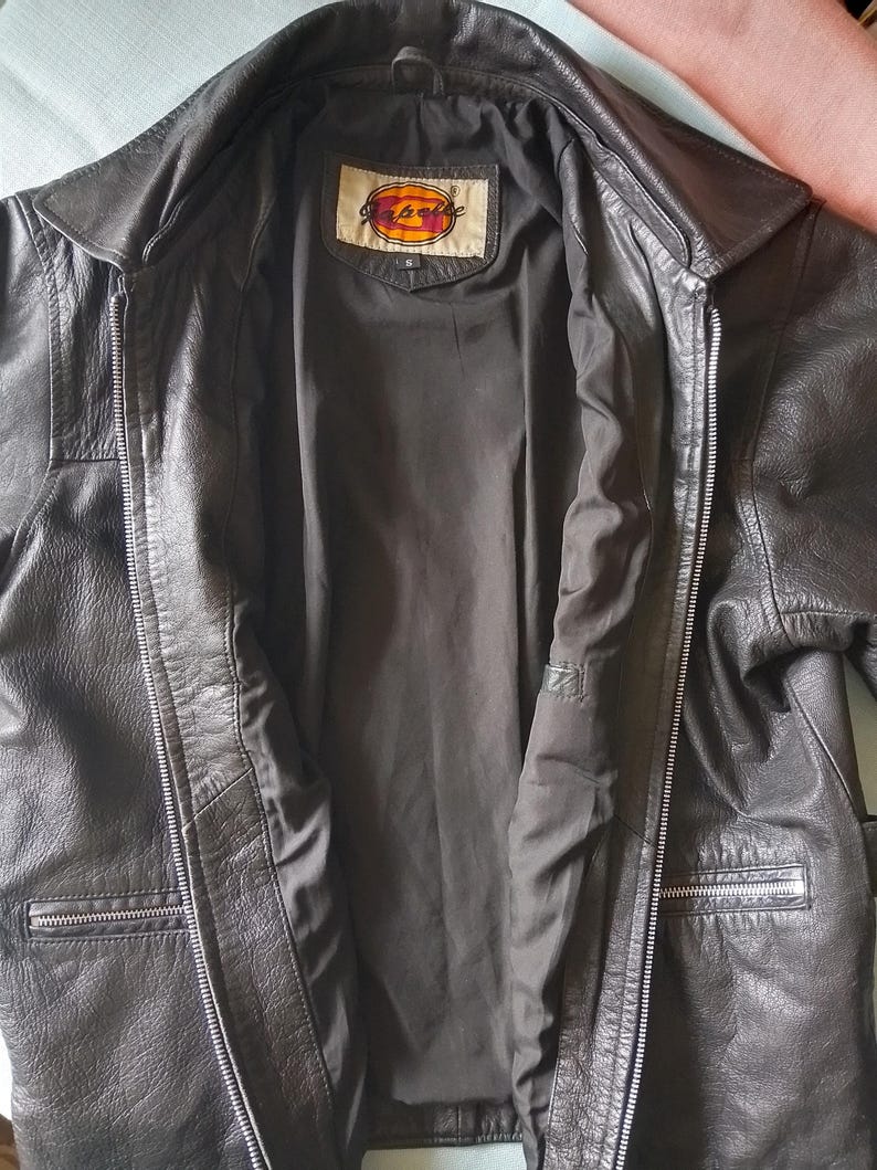 Pu&ograve; includere: Giacca in pelle nera con chiusura a zip e due tasche anteriori. La giacca ha un collo e un'etichetta all'interno del colletto. La giacca &egrave; aperta, rivelando una fodera scura.