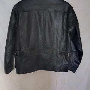 Pu&ograve; includere: Giacca in pelle nera con colletto, cuciture a vista e due cinturini regolabili con fibbia in vita. La giacca ha un design classico e un aspetto strutturato.