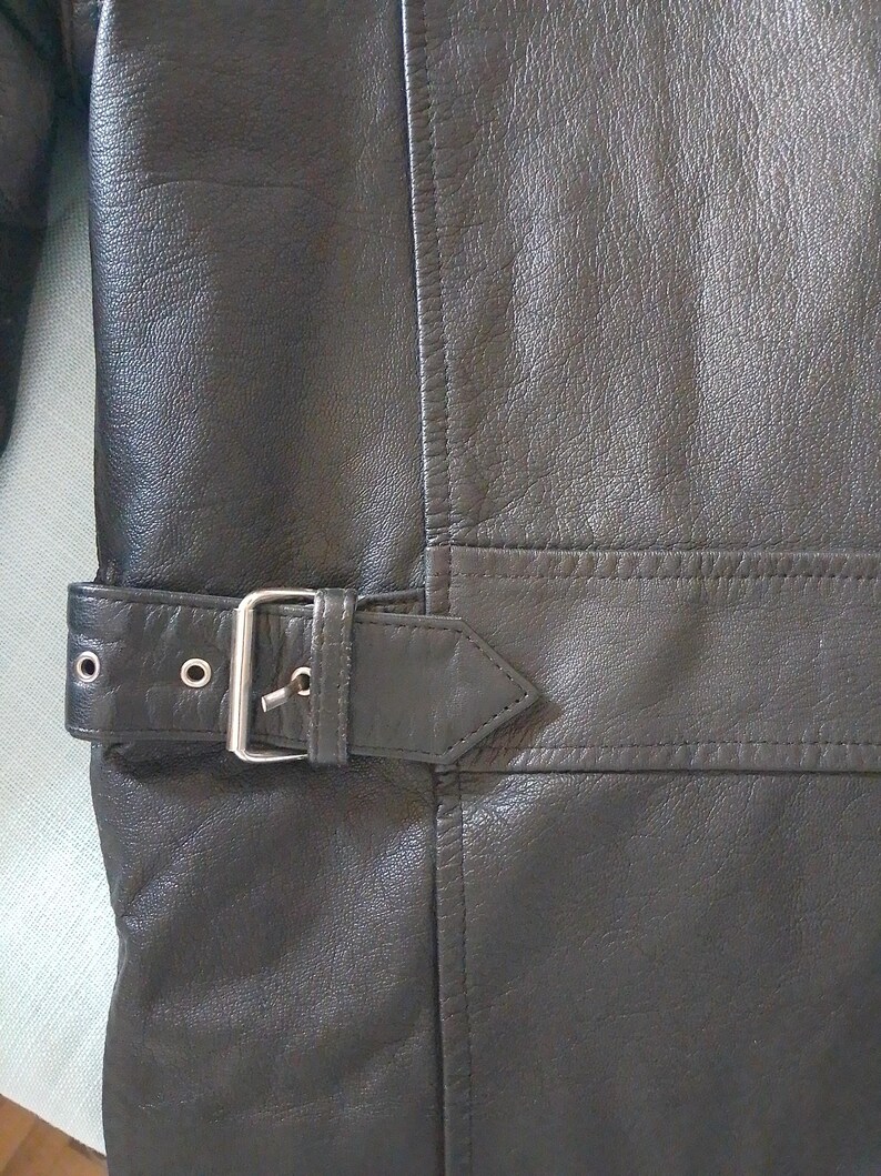 Pu&ograve; includere: Primo piano di una giacca in pelle nera con chiusura a fibbia. La giacca ha una superficie strutturata e dettagli di cucitura visibili. La fibbia &egrave; color argento.