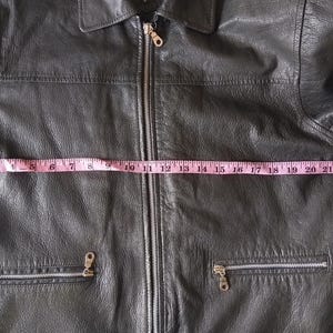 Pu&ograve; includere: Giacca in pelle nera con collo e chiusura a zip integrale. La giacca ha due tasche con cerniera. Un metro a nastro rosa mostra una larghezza di circa 56 cm sul davanti.