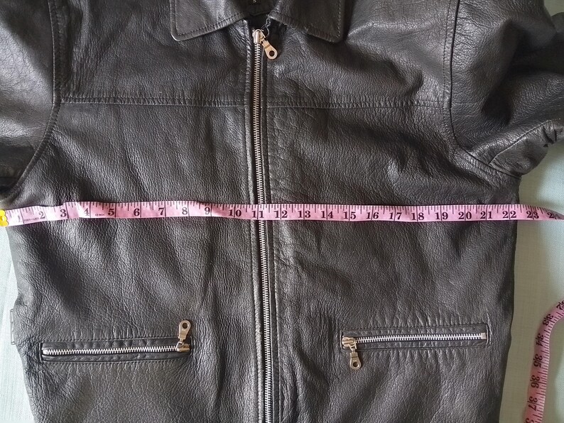 Pu&ograve; includere: Giacca in pelle nera con collo e chiusura a zip integrale. La giacca ha due tasche con cerniera. Un metro a nastro rosa mostra una larghezza di circa 56 cm sul davanti.