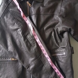 Pu&ograve; includere: Giacca in pelle nera con collo e chiusura a zip. La giacca ha due tasche con cerniera. Un metro a nastro mostra una lunghezza di circa 63,5 cm.