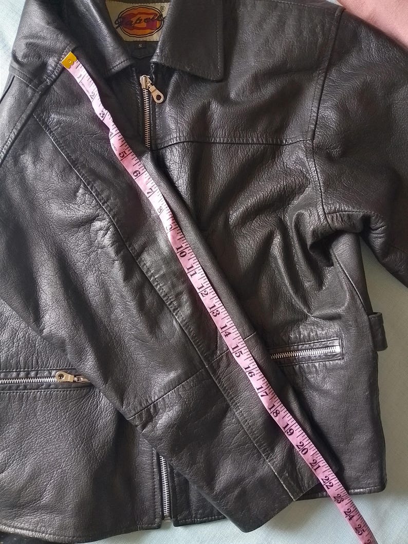 Pu&ograve; includere: Giacca in pelle nera con collo e chiusura a zip. La giacca ha due tasche con cerniera. Un metro a nastro mostra una lunghezza di circa 63,5 cm.