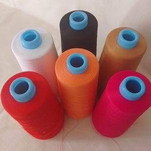 Könnte beinhalten: Sechs Garnrollen in verschiedenen Farben: Rot, Weiß, Schwarz, Orange, Pink und Beige. Jede Rolle hat eine hellblaue Mitte. Die Rollen sind kreisförmig angeordnet.