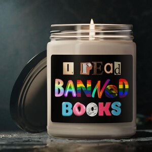 Peut inclure: Une bougie blanche avec une étiquette noire sur laquelle est inscrit "I Read Banned Books". Les lettres sont dans un arc-en-ciel de couleurs. La bougie est allumée et la flamme est visible.