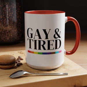 May include: White ceramic mug with a red interior and handle. The mug features the text "GAY & TIRED" in black, with a rainbow-colored stripe below the text. A spoon and a macaron are on a wooden surface.