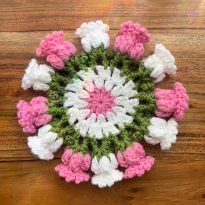 May include: A crocheted coaster with a circular design featuring pink, white, and green flowers. The coaster is made with a variety of stitches and has a textured surface.