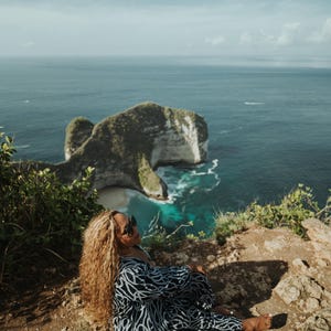 Può includere: Una donna con occhiali da sole è seduta su una scogliera rocciosa che domina un oceano turchese. Un'isola rocciosa verde di grandi dimensioni è visibile in lontananza.