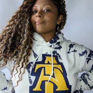 May include: A white tie-dye hoodie with a large navy blue and gold embroidered letter "A" on the front.