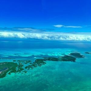 May include: An aerial view of a turquoise ocean with small islands covered in green vegetation. The sky is a bright blue with white clouds.