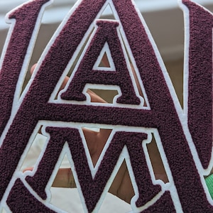 May include: A maroon felt patch with white outlines of the letters "A" and "M".