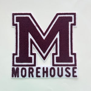 Könnte beinhalten: Ein Patch aus maroonfarbenem Filz mit einem weißen Umriss des Buchstabens "M" und dem Wort "MOREHOUSE" darunter.