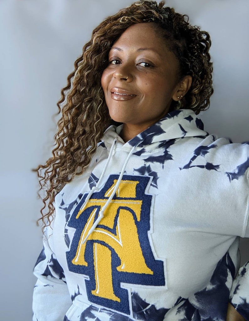 May include: White tie-dye hoodie with a large gold and navy blue "AT" patch on the front.