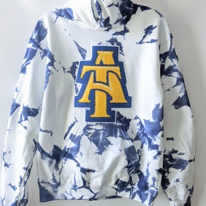 May include: White hoodie with a blue tie-dye pattern and a large yellow and blue "AT" patch on the front.