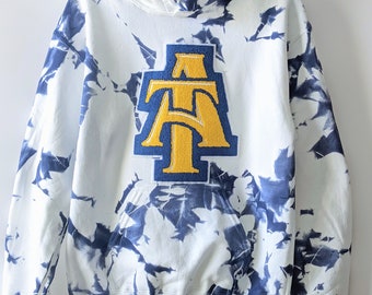 Peut inclure: Sweat-shirt à capuche blanc avec un motif tie-dye bleu et un grand patch "AT" jaune et bleu sur le devant.