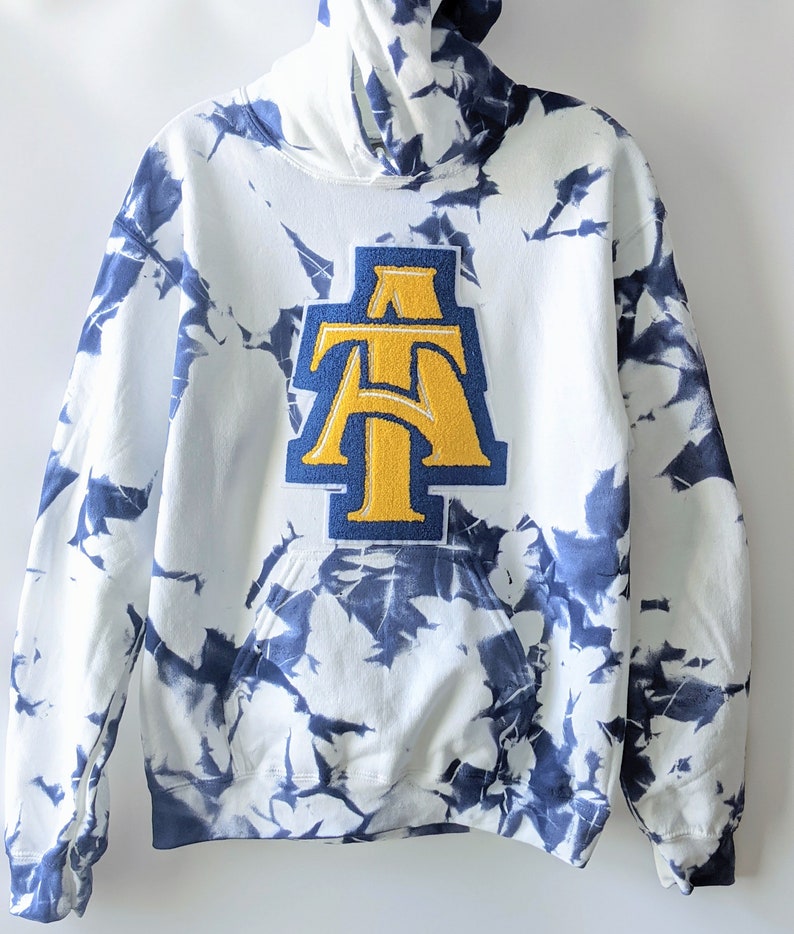 May include: White hoodie with a blue tie-dye pattern and a large yellow and blue "AT" patch on the front.