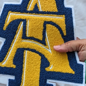 May include: A large, embroidered patch featuring the letters "ATA" in a bold, varsity-style design. The letters are yellow with a navy blue border and white accents, set against a white background. The patch has a textured, chenille-like appearance.