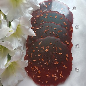 May include: Set of three resin coasters with a dark red base and copper flecks. The coasters are layered and have a unique, organic shape.