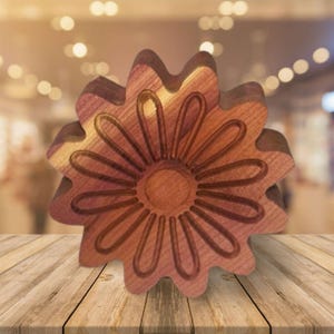 May include: A wooden flower-shaped object with a detailed floral design carved into its surface. The object is made of a light brown wood with a darker brown carved design. The flower has multiple petals and a circular center. The object is sitting on a wooden surface.