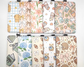 Puede incluir: Una colección de diez portatarjetas rectangulares con varios patrones impresos que incluyen osos, lazos, diseños florales, útiles escolares, balones de fútbol americano y cascos. Cada soporte tiene una anilla de metal en el borde superior.