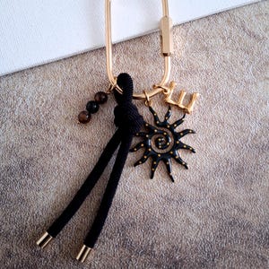 Peut inclure: Porte-clés doré avec un charm en forme de soleil noir, un pompon noir et un petit charm en forme de lettre dorée. Le charm soleil a des accents dorés. Une chaîne de perles marron est également attachée.