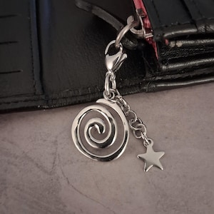 Könnte beinhalten: Ein silberfarbener Anhänger mit einem Spiraldesign und einem kleinen Sternanhänger, der durch eine Kette verbunden ist. Der Anhänger hängt an einer schwarzen Geldbörse mit rosa Innenfutter.